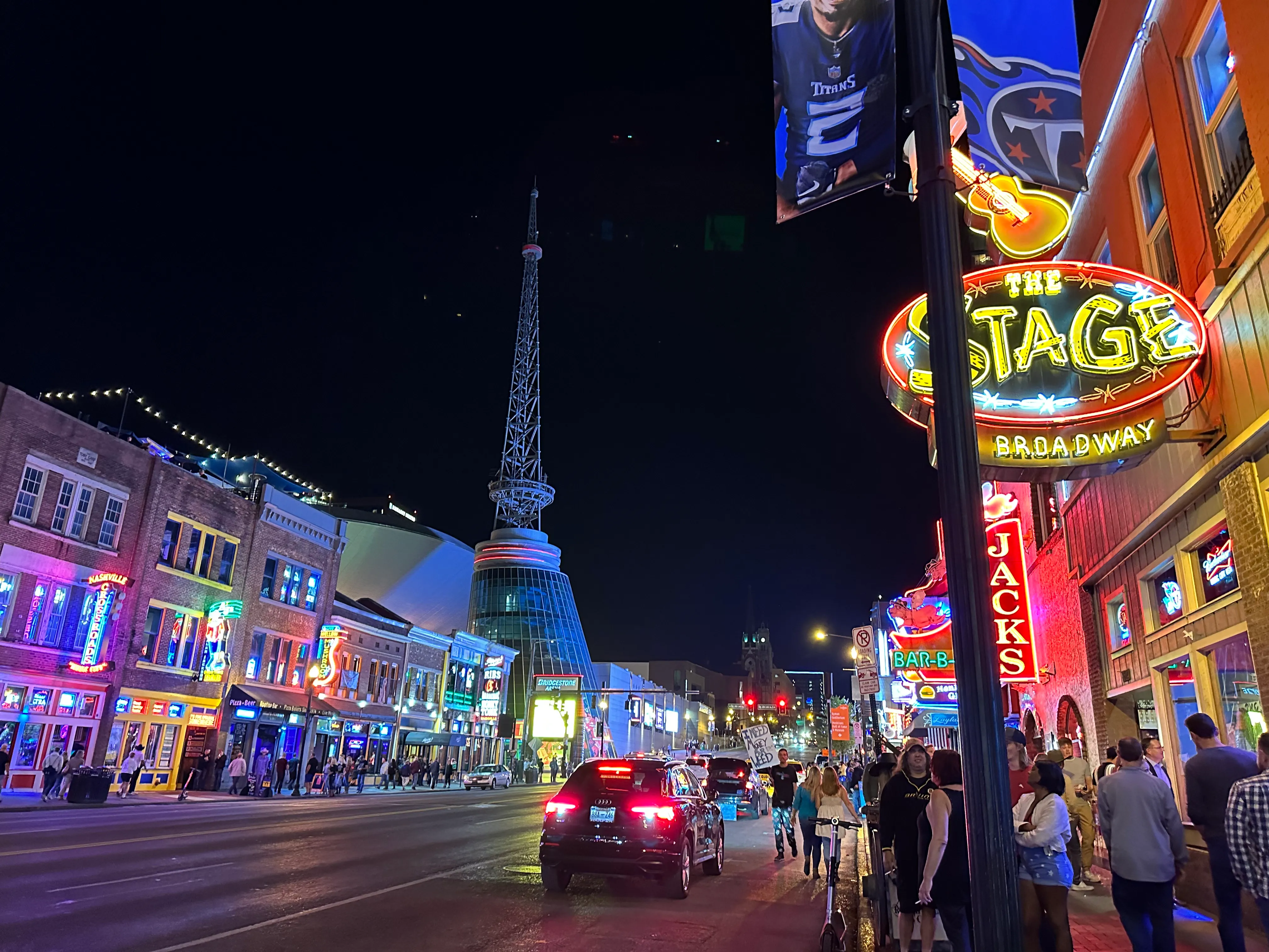 Lower Broadway in Nashville at night: neon signs for The Stage and Jack's Bar-B-Que glow over a busy sidewalk, with the Bridgestone Arena spire lit up down the street.